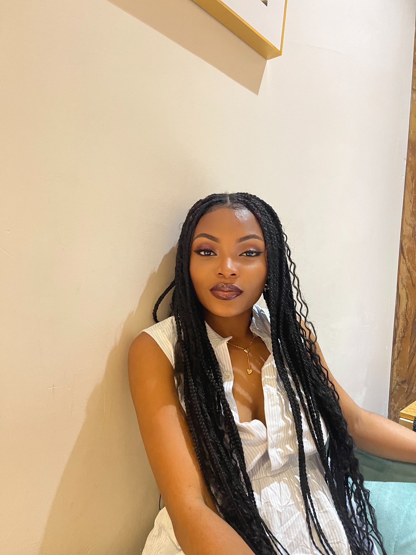 Deborah leaning against a cream wall in a white striped dress with long braids, gold heart pendant necklace, soft daylight.