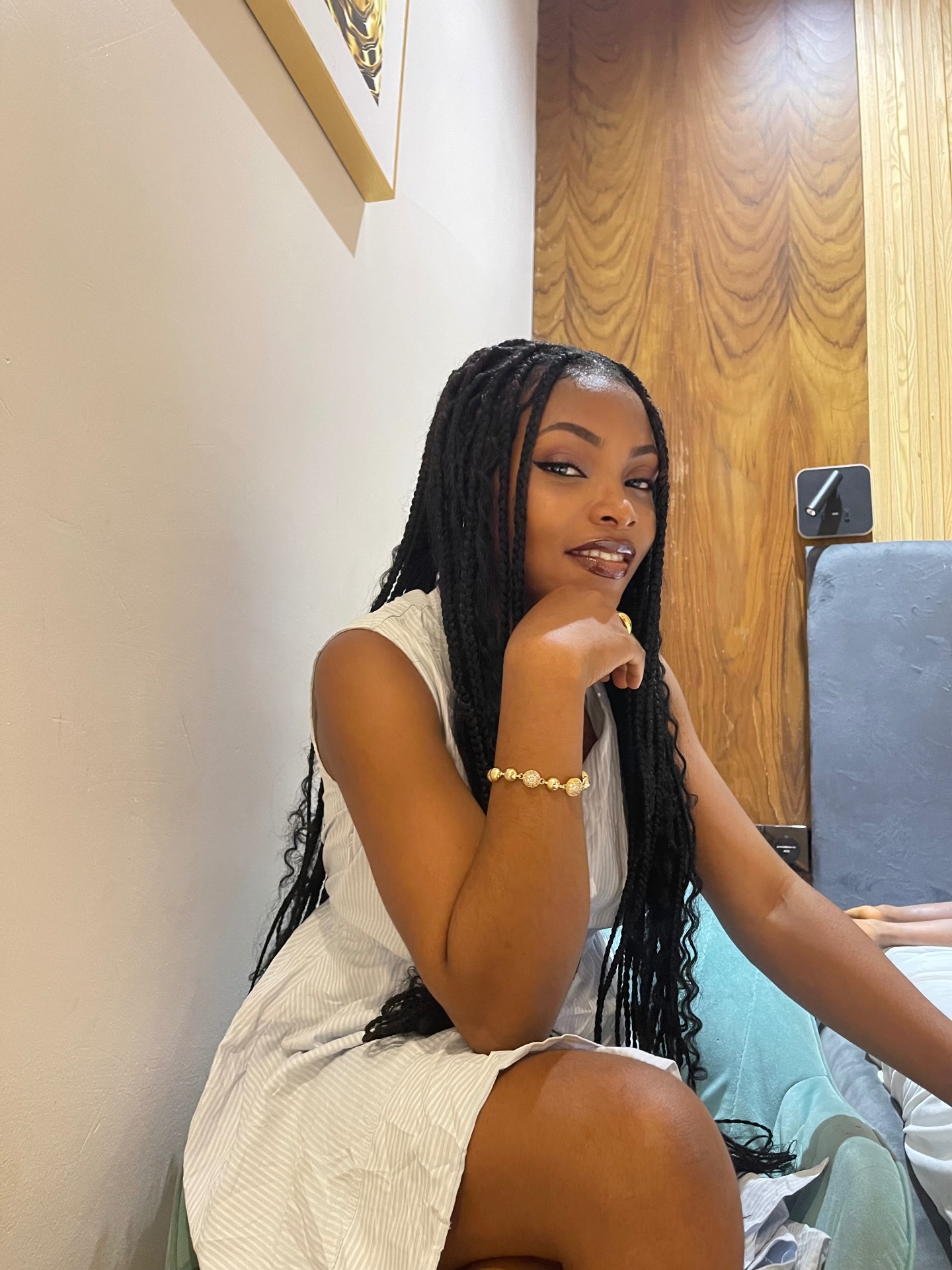 Deborah seated in a white striped sleeveless dress with long braids and a gold beaded bracelet, hand resting under her chin, framed gold artwork on the wall behind her.